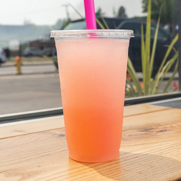 Pink frozen drink in a clear cup with a pink straw on a wooden table by a window.