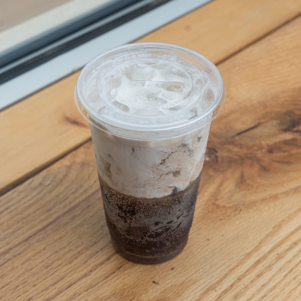 Iced coffee drink with creamy top layer in a plastic cup on a wooden table.