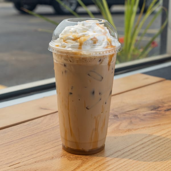 Iced coffee with whipped cream and caramel drizzle on a wooden table by a window.