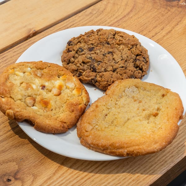 Three cookies: white chocolate macadamia, oatmeal raisin, and sugar, on a white plate atop a wooden table.