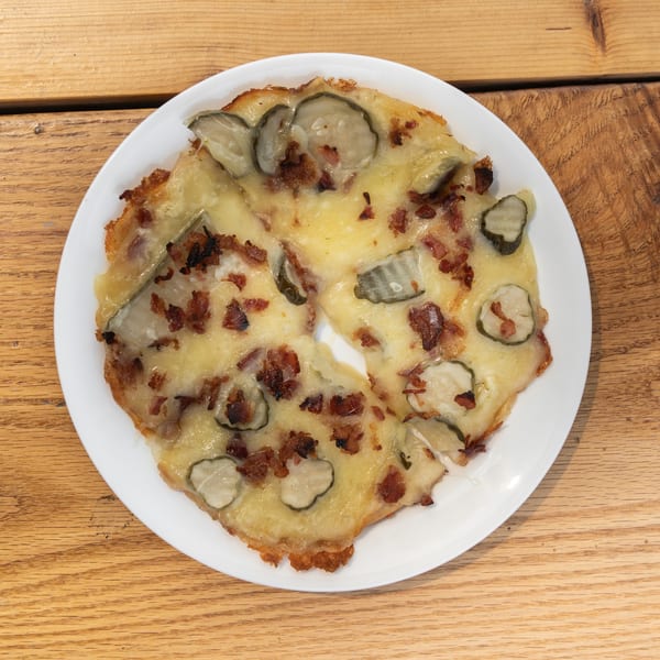 Savory flatbread with melted cheese, crispy bacon, and pickle slices on a white plate.