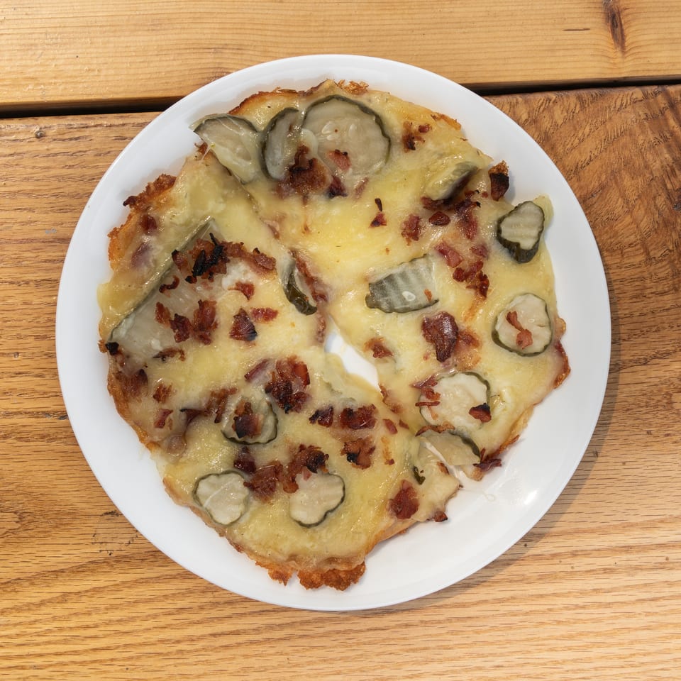 Savory flatbread with melted cheese, crispy bacon, and pickle slices on a white plate.