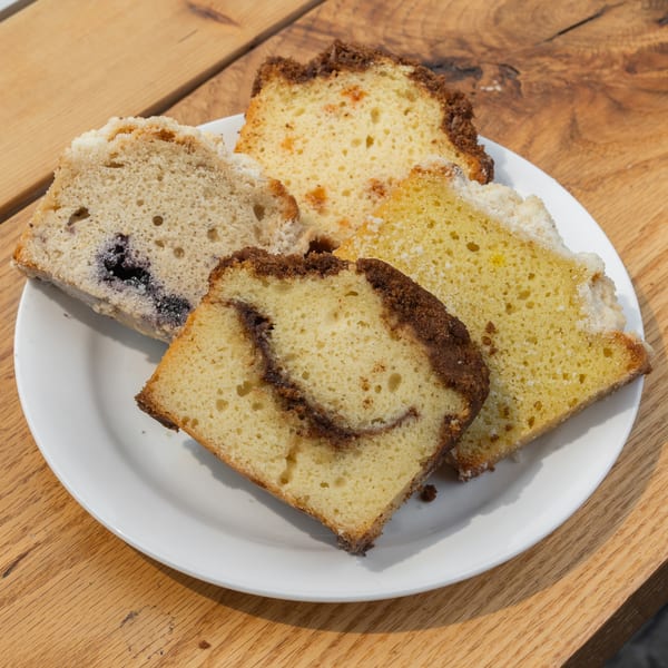 Assorted cake slices, including blueberry, cinnamon swirl, and crumble, on a white plate atop a wooden table.