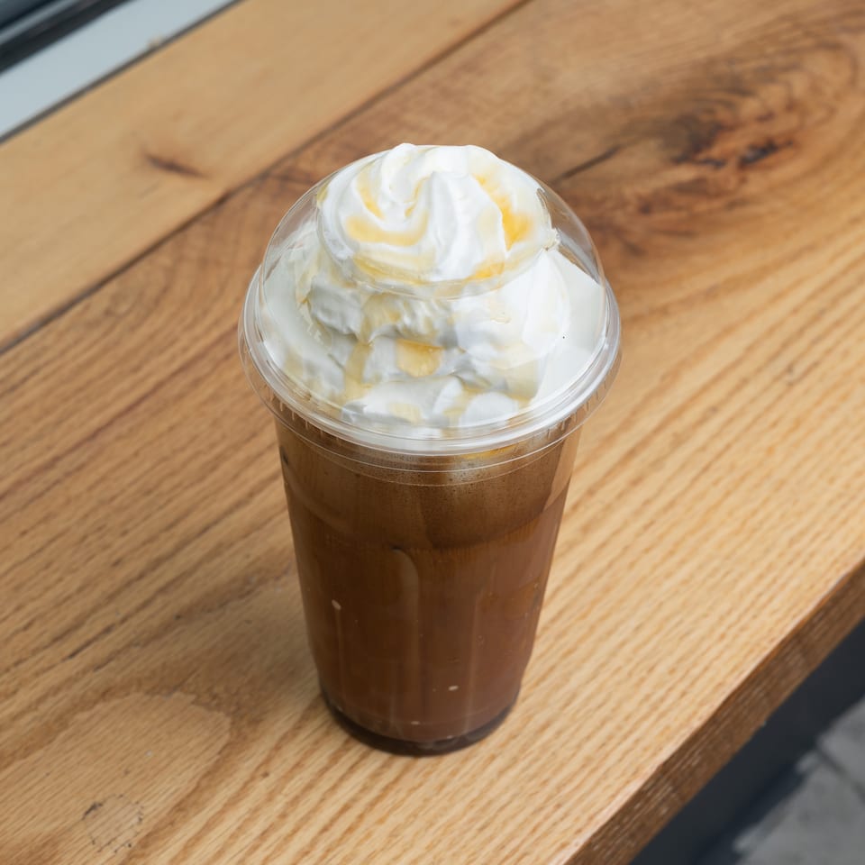 Iced coffee with whipped cream and a golden drizzle in a plastic cup on a wooden surface.
