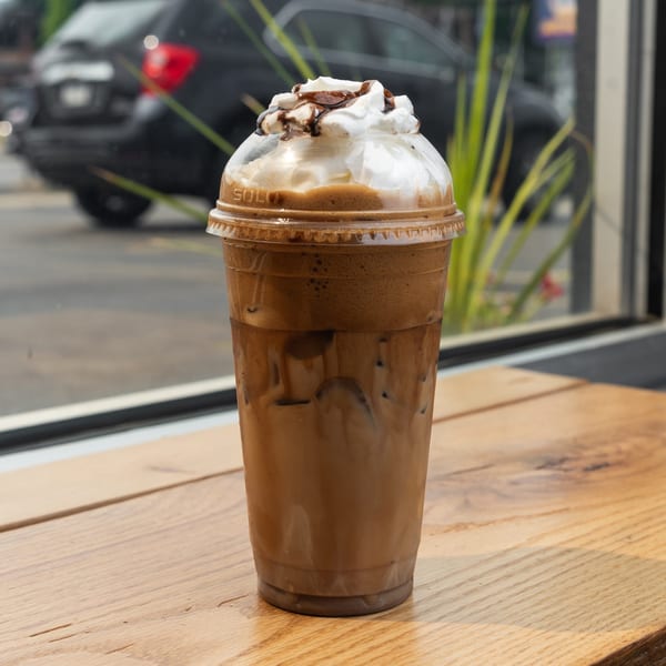 Iced coffee with whipped cream and chocolate drizzle on a wooden table by a window.
